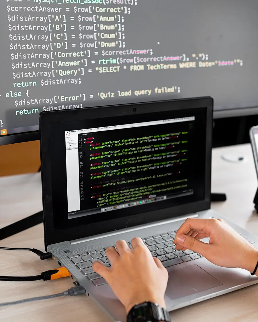 Hands typing on a laptop with code on the screen and a larger monitor behind